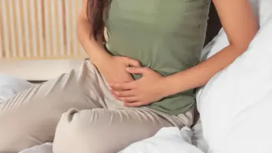 Žena sedí na posteli a drží si břicho kvůli bolesti.