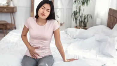 Žena sedí na posteli a drží si bolavý podbřišek.