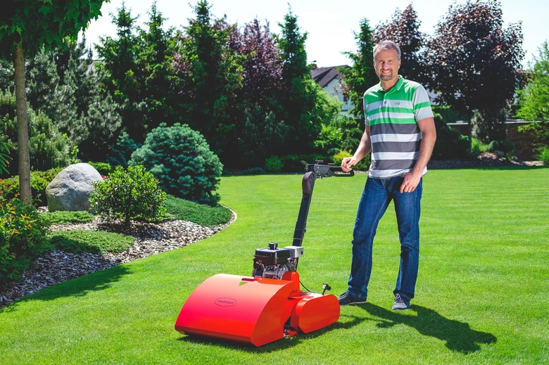 Muž stojí na zeleném trávníku s oranžovou sekačkou.